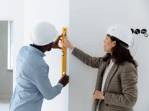 Two people in hard hats checking a wall with a level in a bright room