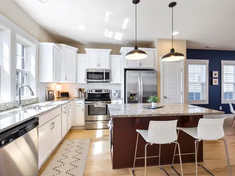 A sunny kitchen with white cabinets, a granite island and white stools
