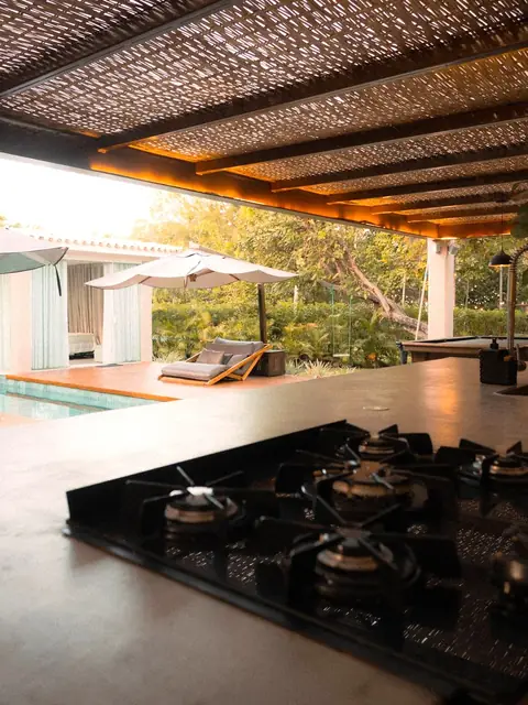 A gas cooktop set in a stone outdoor counter under a reed pergola beside a pool